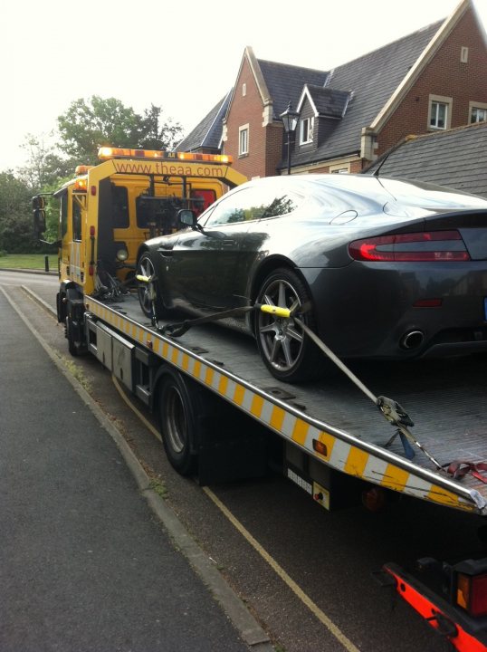 Pistonheads Start Day - The image shows a car securely loaded onto a flatbed tow truck. The car is a convertible, which is partially uncovered due to the open position of its soft top. The tow truck is equipped with a yellow warning light on the top, suggesting it is either passing by or following safety regulations. Surrounding the scene is an urban setting with a road towards the grass on the right side of the frame. There appears to be a residential area in the background with brick houses and a driveway can be seen nearby. A tree is also noticeable in the background, indicating a park or green space close to the residences.