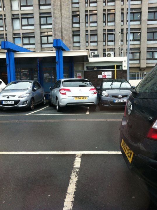 The BAD PARKING thread [vol3] - Page 123 - General Gassing - PistonHeads - The image shows a street scene with several cars parked on the side. The cars are all compact or small models and appear to be in various states of locking or unlocking. Behind the parked cars, there's a building with a distinctive blue facade, indicating no people are present at the moment. The sky is overcast, suggesting a cloudy or possibly rainy day. A line of parked cars forms the foreground of the image, leading from the trash can near the building to where the photo is being taken. The background consists of the building and a stretch of pavement.