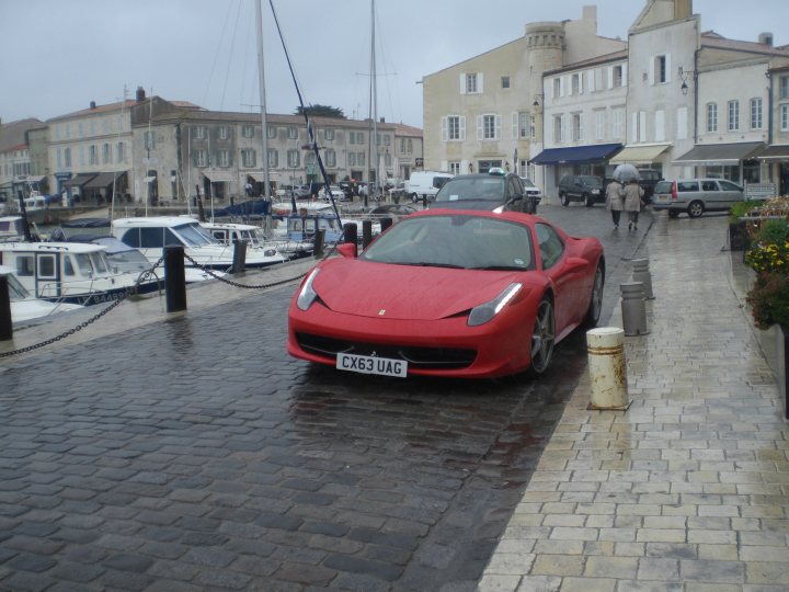 Free car photoshoot - Lynton. Exotica required - Page 1 - South West - PistonHeads - The image presents a serene seaside view. A red Ferrari car, sporting the tag CX83 UAL, is parked on a cobblestone street. The street is lined with buildings and boats are visible in the background, nestled in a marina. The image gives off a European vibe with its brick roadway and nautical elements. A few potted plants add a touch of nature to the scene. The Ferrari's vibrant red color contrasts beautifully with the muted tones of the cobblestone street.