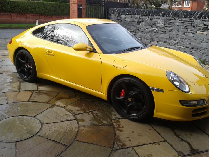 Yellow porn - Page 1 - 911/Carrera GT - PistonHeads - The image shows a yellow Porsche sports car parked on a driveway. The car has two doors and is characterized by its sleek design and large air intakes. The paint appears to be of a high-quality finish, and the trim around the windows is black, providing a nice contrast against the yellow body of the car. There is no text visible in the image, and the focus is solely on the vehicle. The setting seems to be in a residential area, given the style of the driveway and the presence of a home in the background. The car seems to be parked neatly on the driveway, which is made up of interlocking stone tiles. The backdrop features a stone wall, which adds a classical touch to the scene.