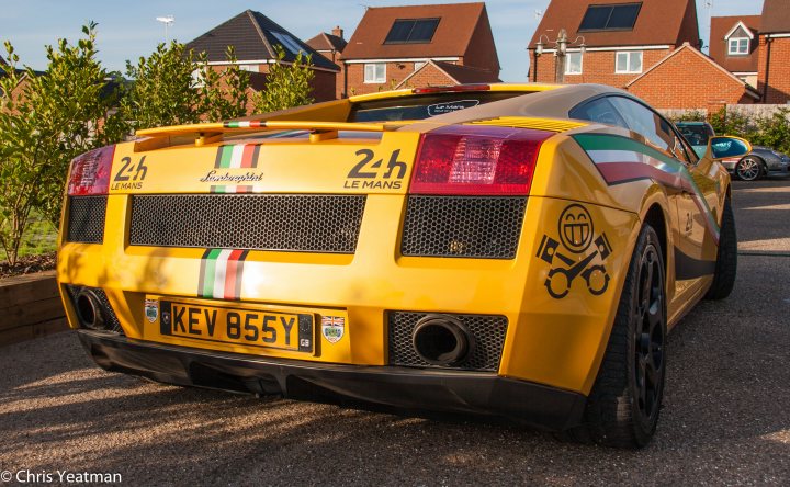 A red double decker bus parked in a parking lot - Pistonheads - The image features a bright yellow sports car parked on a sunlit gravel driveway. The car boasts a yellow rear wing, a prominent 'Mazda' logo on the front, and a license plate that reads 'K4V 855Y'. The tail lights indicate that it's a high-performance quadrot Letour-Le Mans, and Italy is specified with a license plate mention. The car is set against a backdrop of red brick houses with brown roofs, adding a quaint residential feel to the scene.