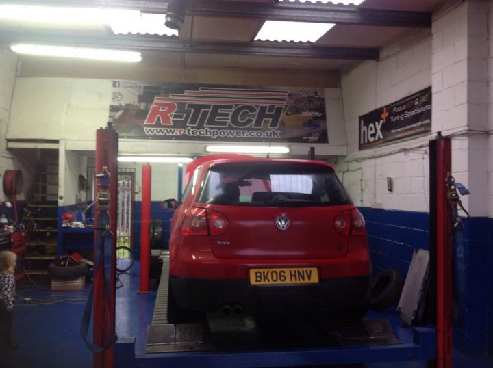 Golf GTI DSG in Red! - Page 2 - Readers' Cars - PistonHeads - This is a photograph taken in a workshop garage. Amongst various vehicles and equipment, one red car is hoisted on a lift, suggesting maintenance or repair work. The setting includes a banner promoting the local services available at the garage. There is a person visible at the floor left of the frame and safety gear like a fire extinguisher.