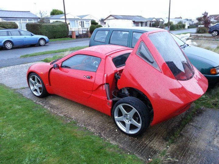 Dragfepic.pw - The image shows a vibrant red sports car parked on a brick pavement next to a grassy area. The car's design is sleek, with an open driver's compartment exposing its engine and gearbox, giving it a unique and unusual appearance. Another car is partially visible in the background, parked further away. The weather appears to be overcast, and there are residential buildings in the distance. The photo gives a sense of discovery due to the unusual positioning of the car.