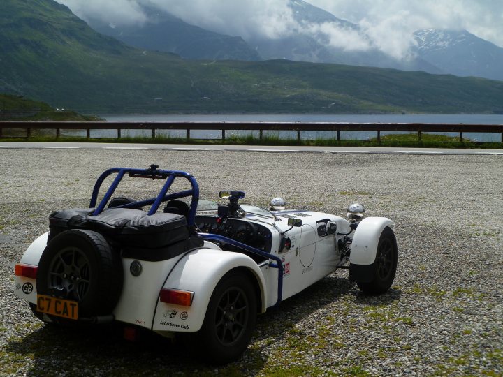 Euro trip 7 vs vx220 - Page 1 - Caterham - PistonHeads - The image captures a moment of tranquility in a mountainous area, where a small sports car with an open roof and two prominent side vents is parked on a gravel lot, overlooking a serene lake nestled between the towering green mountains in the background. The car's position suggests it has stopped to take in the picturesque view. The surrounding greenery complements the natural colors of the landscape, and the overall scene speaks to the serenity and beauty of nature.