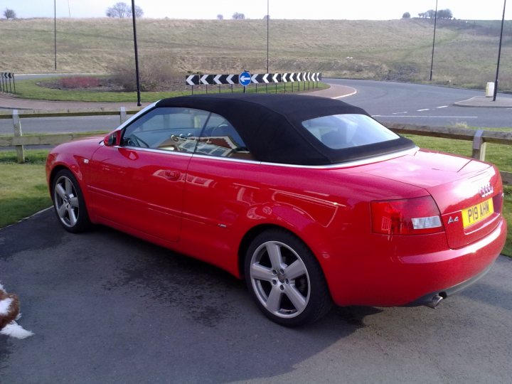 My Car - Page 1 - Readers' Cars - PistonHeads - The image displays a red convertible car parked on the side of a road. A detachable black soft top is folded back, revealing the car's interior. The setting is an open-ground area with a grassy field in the background, and it appears to be in a rural or less urbanized environment. In the foreground, there is a section of asphalt leading to a roundabout, indicating a concrete surface. There are no texts, people, or other noticeable objects in the immediate vicinity of the car. The sky above is overcast with no distinct features.