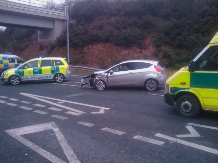 Show us your crash pics!! - Page 66 - General Gassing - PistonHeads - A traffic accident scene is visible, with several vehicles including an ambulance involved. One of the cars, a silver hatchback, is marked with significant damage with a broken windshield and visible scratches and crumples along the front and side. An orange and white ambulance is positioned to the left of the damaged car, and a green and yellow vehicle is on the opposite side of the accident scene. The road is a two-way highway, and the vehicles appear to be moving away from the accident towards a reddish hill. The day is overcast with no visible signs of people in the immediate vicinity of the accident.