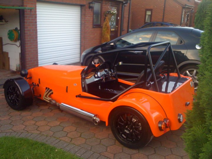 Contribution Pistonheads - The image depicts a vibrant and unique-looking orange Volkswagen Beetle-style car parked in a driveway. The car's color stands out against the muted tones of the surroundings. The vehicle appears to be in good condition, suggesting it may be a collectible or well-maintained. In the background, there is a person who appears to be exiting or entering the car, as well as another car partially visible on the right side of the frame. The style of the image seems informal and possibly taken with a smartphone or over a casual walk.