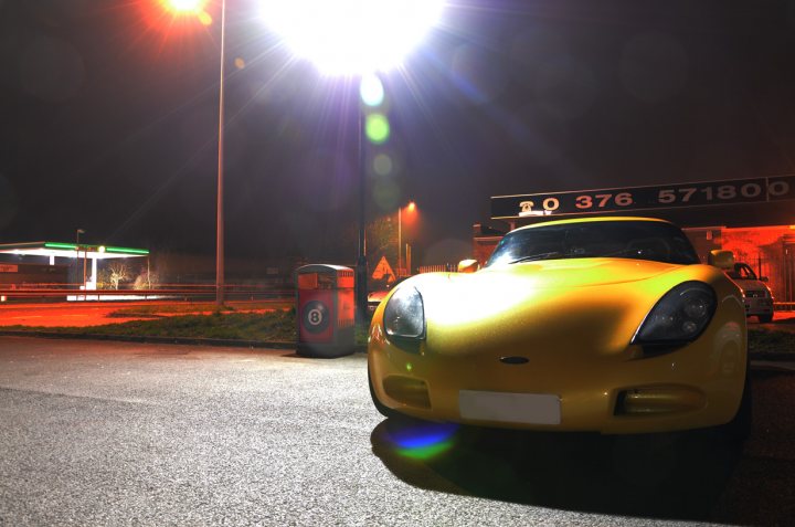 March Essex Pistonheads - The image presents a vibrant night scene featuring a striking yellow sports car. The car, associated with "Everyman's Tennis Club", is parked in an empty lot. It's a miniature-sized sports car, adding a sense of whimsy to the image. A gas station is visible in the background, underscoring the nocturnal setting. The car's metallic sheen is captured under a streetlight, casting a warm glow and adding depth to the image.