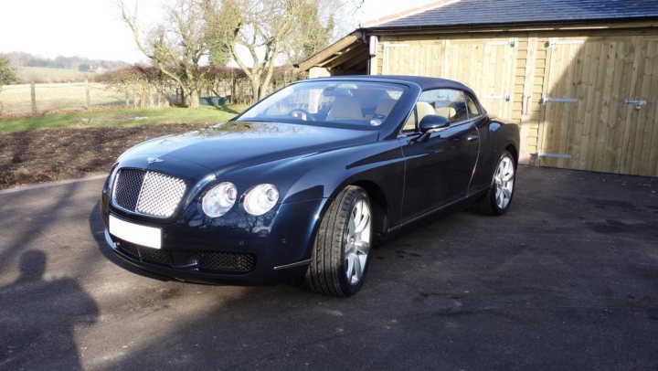 A car parked in a parking lot next to a car - Pistonheads - The image shows a black Bentley vehicle parked in a driveway beside a wooden structure, which resembles a garage with multiple doors. The car appears to be a convertible model with a sleek design, featuring characteristics like round headlights, a chrome grille, and alloy wheels. There is no visible text in the image, and the background suggests a rural or semi-rural setting with greenery and grass indicating a garden area. The lighting and shadows suggest it is daytime.