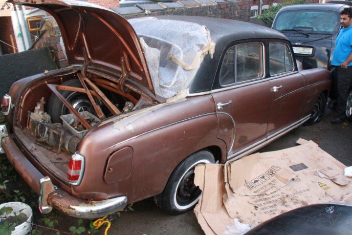 1950's Merc 220s Saved from the grave - rat rod restoration  - Page 1 - Mercedes - PistonHeads - The image shows an older model car being lifted into the air using a hydraulic dead car puller. The vehicle appears to be a brown sedan with an open trunk. The trunk's interior is revealed and is filled with spare parts and tools. A man is standing next to the vehicle, observing the process of the car being lifted. The car is placed on a piece of plywood, which is laid out on the ground below it.