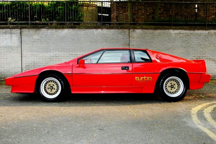 Pick Late Pistonheads - The image features a vibrant red classic sports car parked on an urban street. The car stands out due to its striking color and its position in front of a white brick wall, creating a bold contrast. The vehicle belongs to the BURT group, as indicated by the yellow text on its side. The car's vintage design and large rear wing, a characteristic feature of racing cars, hint at its speed capabilities. Despite being a still image, one can almost feel the raw power it could deliver when it comes to life on the roads.