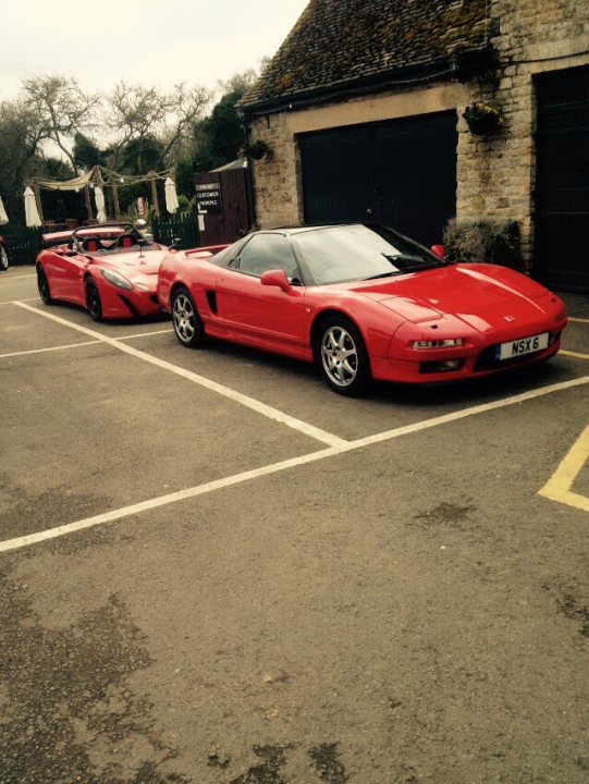 Cars around the Green - Evenley South Northants - Page 21 - Northamptonshire - PistonHeads - The image captures a scene of luxury and speed, featuring a striking red Ferrari parked in a parking lot. The Ferrari is positioned in the middle of the image, drawing the viewer's attention with its distinct design and vibrant color. The other car, also red but without the characteristic Ferrari design, is parked nearby, adding a sense of balance to the scene. The parking lot is edged by a wooden fence and brick buildings, providing a contrasting backdrop to the modernity of the Ferraris. Despite the emptiness of the setting, the presence of multiple umbrellas suggests a recent event. The scene is tranquil yet holds the promise of action and movement.