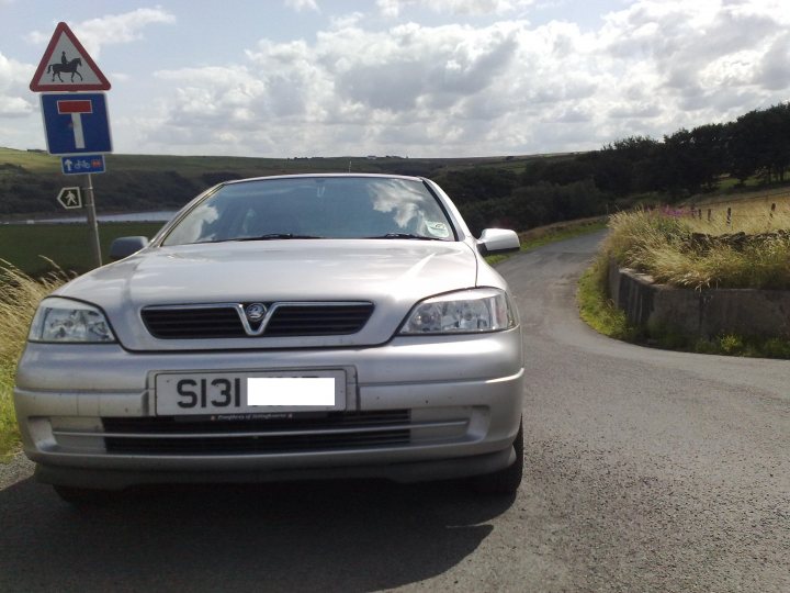 Pistonheads - In the image, a silver Vauxhall car is parked on the right side of a winding, country road. The car is facing towards the left of the frame. On the left, there is a traffic sign, painted in blue and red, indicating a T-junction ahead. The road is nestled among green hills, adding a sense of tranquility to the scene.