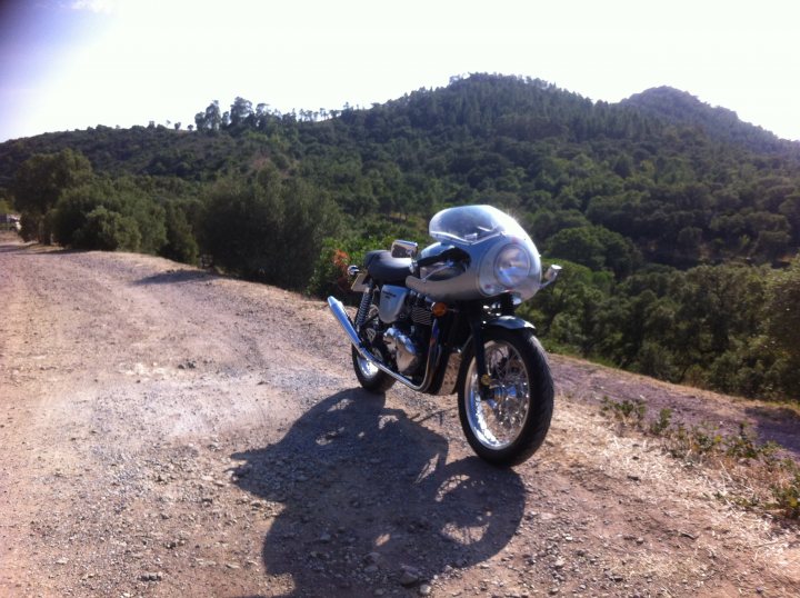 Driving France - Page 4 - Chimaera - PistonHeads - The image shows a motorcycle parked on a gravel road, facing towards the camera. The motorcycle appears to be well-maintained and of a sporty design. Behind the motorcycle, the landscape is lush with green trees and bushes, hinting at a rural or countryside setting. In the distance, there is a clear view of a hill with sparse vegetation, suggesting a somewhat arid environment. The sky above is mostly cloudless, indicating fair weather. The scene promotes a sense of solitude and an appreciation for the outdoors. There are no visible textures or distinctive logo patterns, keeping the focus on the motorcycle and its surroundings.