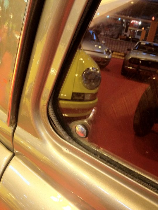 Renault Gordini Dauphine 610 1960 Newbie - Page 1 - French Bred - PistonHeads - The image captures a close-up view of a car's interior, focusing on the sun-filtering rear view mirror. The mirror is rectangular and tinted a light maroon hue, reflecting the dashboard of the vehicle from which the image is taken. The mirror's reflection shows that the car is parked indoors, possibly in a showroom or dealership, given the presence of other cars visible in the background. The ambient light seems to be coming from the top left corner, suggesting a natural light source. The mirror's transparent base suggests a modern vehicle interior design.