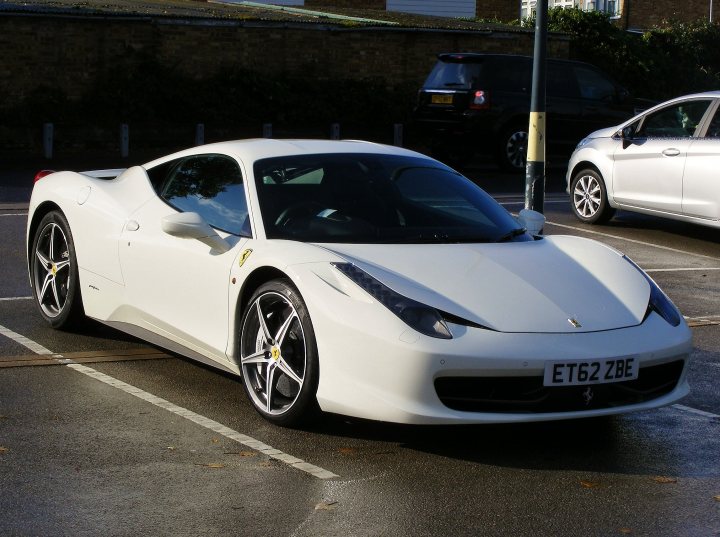 A car is parked on the side of the road - Pistonheads - The image displays a white sports car, specifically a Ferrari, with a black license plate, parked in a parking lot. The car has the iconic yellow Ferrari emblem on the side and features a sleek design with a low-profile appearance. It is parked near other cars, and the setting suggests a residential or urban environment with brick buildings. The sun is shining on the car, highlighting its white color.