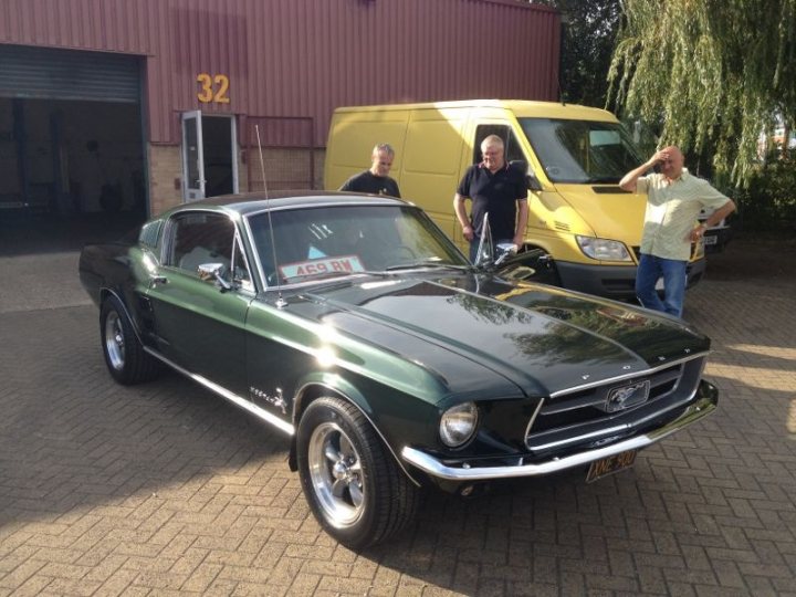 Wheeler Dealers - New Series 2012 - Page 16 - TV, Film & Radio - PistonHeads - The image is a photograph that depicts a vintage Ford Mustang parked next to a large yellow van and two men standing between them. The car is positioned facing left, its side profile highlighted against the more modest vehicle next to it. The setting appears to be a paved area, possibly a parking lot, with tall buildings in the background, suggesting an urban environment. One of the men is posing near the front of the car, while the other stands slightly behind them. Both men are dressed casually, and the bright sunlight suggests it's daytime.