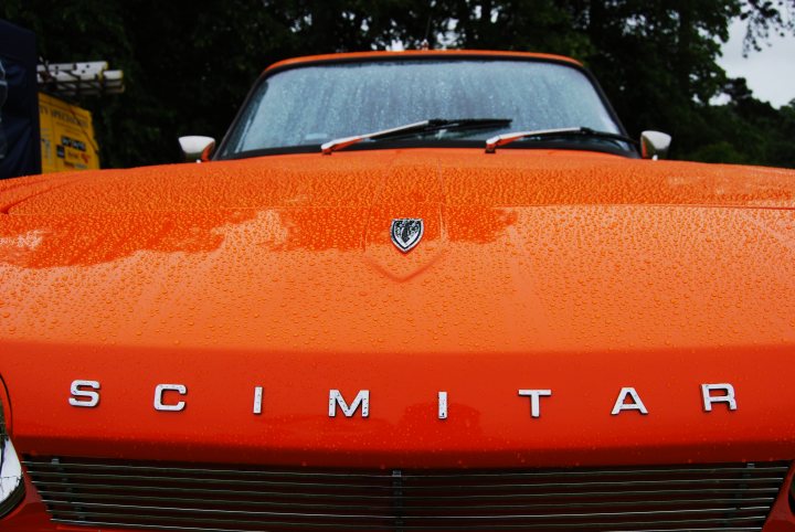 A red car is parked in a parking lot - Pistonheads - The image shows a vibrant, retro-style convertible car. The car's hood has numerous water droplets scattered across it, suggesting recent rain. The color of the car is a bright orange, and it features a prominent silver emblem in the center of the hood. The vehicle also has a unique silver on the side of the hood. The surrounding environment includes trees, indicating that the photo might have been taken outdoors, possibly in a park or a forested area.