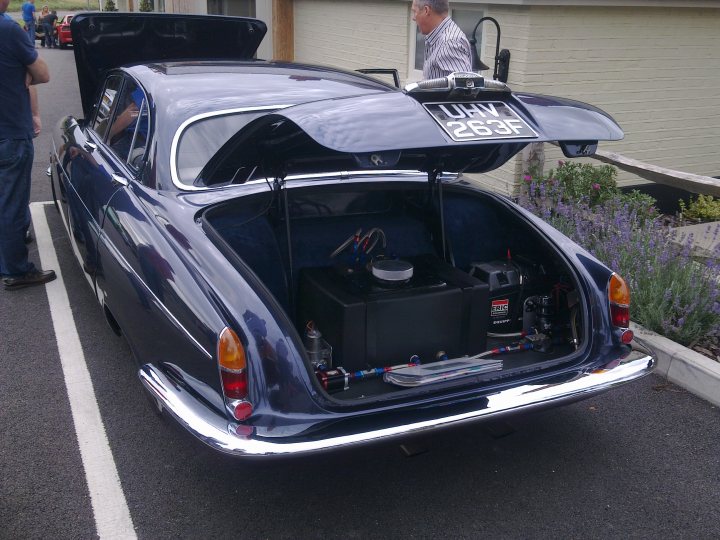 Pistonheads - The image shows a vintage car with its trunk opened, revealing a variety of items inside. The car is parked on the side of a road, near a building. A person is standing next to the car, observing the trunk's contents. The license plate on the trunk reads "LBW 126". There is also a motorcycle in the background. The number "265" is visible on the car. The scene appears to take place during the day under clear skies.