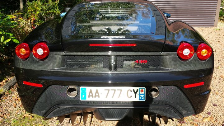 New Scuderia Owner - What a car!!! - Page 1 - Ferrari V8 - PistonHeads - This image captures a black Ferrari sports car situated on a gravel surface. The vehicle appears to be a limited-edition model, indicated by the words "AA-77-CY" on the license plate, which is prominently displayed in the foreground of the image. The Ferrari is topped with a black convertible roof, providing a contrast with the car's overall black theme. The car's position on a gravel driveway or similar surface, together with the convertible top, suggests a setting possibly aimed at displaying the vehicle. The license plate itself becomes a central point of interest within this particular context.