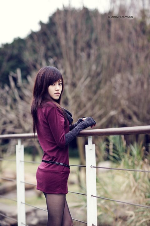 This image seems to capture a moment of tranquility. A young woman, styled in dark purple, stands confidently with a slight lean against a wooden railing. Her gaze is directed off to the side, her expression thoughtful. Behind her, the soft focus of a black and white photo adds a retro feel, with trees hinting at a natural outdoor setting.