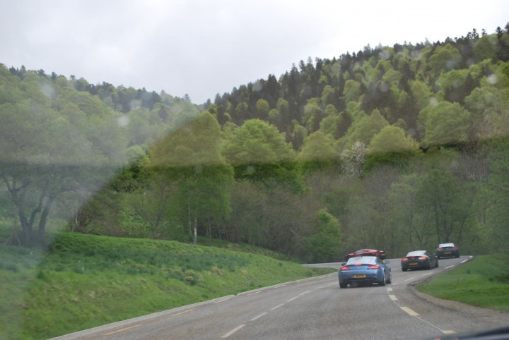 Pistonheads trip to Monaco Historique - Page 2 - Aston Martin - PistonHeads - The image shows a winding road through an area of lush green trees. There are at least three cars on the road, suggesting a traffic situation or perhaps a holiday drive if it's during a time of high travel. The sky is overcast, casting soft light on the scene. The photograph has a naturalistic quality, as if taken from the perspective of someone inside a car, with the windshield streaked by rain or wet from wiper use.