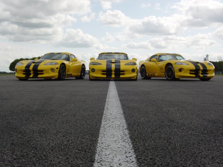 The image showcases three vibrant yellow race cars parked in a straight line on a tarmac introduction area. Each car is unique, with one featuring wide tires and another showcasing a distinctive black and yellow livery pattern. They are basking in natural light with clear skies, implying they are outside during the day. The cars are positioned in the center of a wide street, with the one on the left slightly offset. The perspective of the photo suggests it was taken from behind the cars, giving a clear view of their designs and colors.