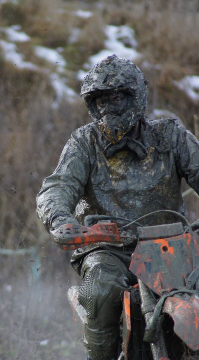 Pistonheads Wild Day Woolly Boxing - The image depicts a person wearing a dark camouflage suit that appears to be covered in dirt or mud. The individual appears to be riding a motocross bike, as suggested by the presence of visible dirt on the bike as well. The person is in the midst of a forested area, with trees and greenery suggesting an outdoor, natural setting. There seems to be a slight blur to the image, which may indicate movement or shake from the bike.