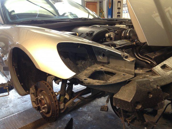 Re-installing the side marker lamps on a UK registered C6? - Page 1 - Corvettes - PistonHeads - The image shows the undercarriage of a silver car being serviced or modified. The vehicle is lifted and supported on black jack stands, indicating that it is stationary to perform maintenance. The front right wheel is visible and seems to be unattached, remaining in its usual position. The presence of parts on the ground and a person's foot visible close to the car signifies an active setting. The car is potentially a modern, high-end model, as suggested by the silver paint and the fluorescent garage environment.