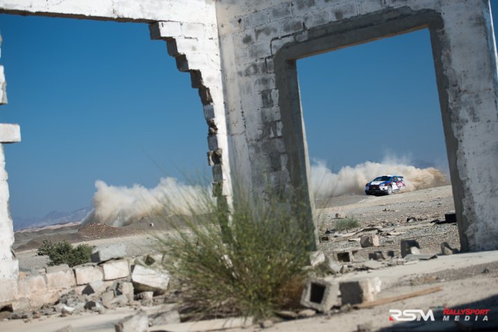 My Photos of the Year - Page 1 - Middle East - PistonHeads - The image depicts an exhilarating scene of a rally racing car navigating through a rough terrain path cut through stone ruins. The car, identified by its striped pattern, is seen mid-action, kicking up dust due to the car's high speed. To the left of the frame, a partially damaged brick wall provides context to the car's surroundings, while on the right side, a hole in the wall opens up to a clear blue sky, providing a stark contrast to the rugged landscape. The dense vegetation suggests a desert environment, which supports the dynamic nature of the scene. The photo is taken from a distance and offers a broad view of the entire course, emphasizing the sport's struggle against nature's elements.