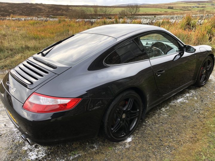 Used 997s - How low do they go and what's selling? - Page 3 - 911/Carrera GT - PistonHeads - The image shows a black Porsche sports car parked on a gravel surface. The vehicle has a sleek design with aerodynamic bodywork, a large rear spoiler, and a low profile. It features a sunroof, custom wheels, and tinted windows. The Porsche logo is visible on the hood and side of the car. In the background, there's a lush green landscape with trees under a partly cloudy sky, suggesting a rural or semi-rural setting.