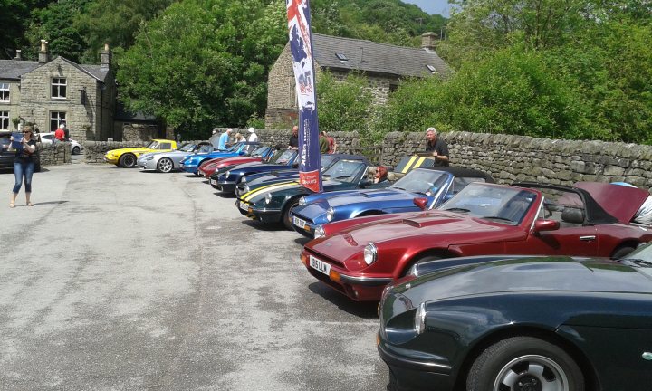 S Club Barn Farm Camping 2016 - Page 6 - S Series - PistonHeads - The image displays a lineup of vintage cars parked in an orderly manner. The cars vary in design, suggesting a diversity of models. The vehicles are parked alongside a grassy area and a stone wall, creating a contrast with the urban environment. A handful of people are scattered around the area, possibly engaged in socializing or admiring the cars. A red and blue banner hangs above the cars, possibly indicating a special event or gathering related to the vintage vehicles.