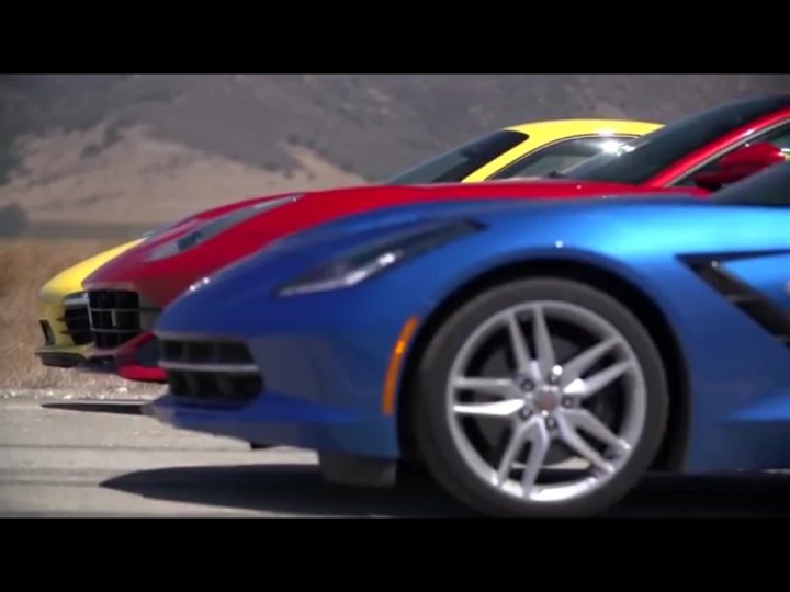 Corvette C7 STINGRAY vs. Porsche 991 vs. Ferrari F12 - Page 1 - Corvettes - PistonHeads - The image shows three sports cars parked next to each other on a concrete surface with blurred mountains in the background. The cars are sleek and angular with large, aggressive grilles, a notable design feature of performance vehicles. The color scheme of the cars includes shades of blue, red, and yellow, adding contrast and speed to the scene. The reflection and positioning suggest that these cars are the focus of the image, and that the environment is currently calm, with no visible movement or activity. The image also includes a watermark indicating that it is from a film or video segment.