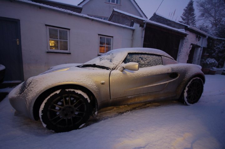 Snow Lotus Pistonheads - The image depicts a silver convertible streetcar parked in winter conditions. The car is covered with a layer of snow, indicating recent snowfall. The streetcar appears to be stationary, with its roof and windows dusted with snow. It is situated in front of a house with a white facade, suggesting it's a residential area. The time is evening, with dim lighting illuminating the scene, giving a tranquil and snowy ambiance to the photo.