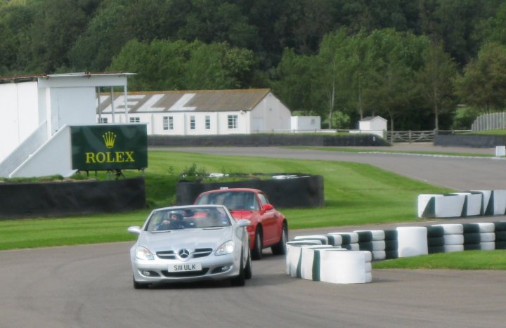 25th august Trackday - Page 1 - Goodwood Events - PistonHeads - This image depicts a silver car making a turn on a road. The car is driving alongside a red car. A grassy area flanks the street, and in the distance, there's a sign for a Rolex store, indicating that this image might have been taken near a racing circuit, considering the presence of a zip tying around the cone course poles. The weather appears to be sunny, and the overall scene suggests a typical day at a racing facility or on a well-maintained public road.