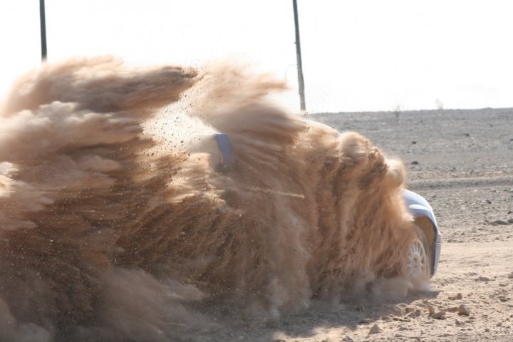 Dubai Rally - Page 1 - Middle East - PistonHeads - The image captures a dynamic scene with a car throwing up a cloud of dust. The car appears to be skidding on its side, with the front tire partially buried in the sand. The dust is scattered around it, indicating rapid motion. The landscape in the background is barren and appears to be a field or a desert. The main focus of the image is the front half of the car, emphasizing the intense motion and the rough, dusty conditions.