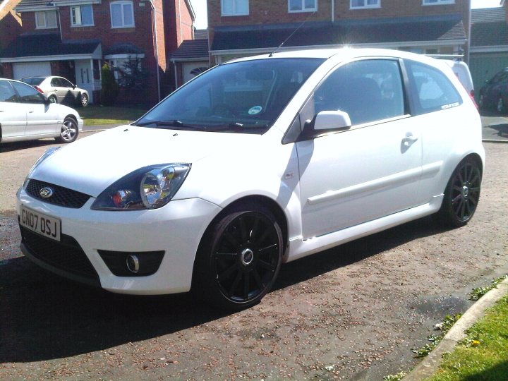 Pistonheads Alloy Refurbs - The image depicts a white hatchback car parked on a street. The car is equipped with black rims and has a visible???rm on the rear window. In the background, there are residential buildings, suggesting a suburban setting. Additionally, there are other cars parked further away. The sky is partially visible, indicating it might be a sunny day.