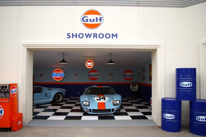 2009 Revival pictures - Page 6 - Goodwood Events - PistonHeads - This image depicts an interior view of a car showroom or a garage dedicated to Gulf Racing cars. The floor is tiled in a semi-circle pattern with alternating black and white. There are several orange soda vending machines to the left, some of which appear to be empty. Two vintage Gulf racing cars, specifically replicas, are parked in the room, one with the number 13 on it and the other with the number 6. The walls are adorned with various logos, predominantly featuring the 'Gulf' brand, creating a branded and themed environment. On both sides of the entrance, there are stacks of blue barrels, possibly used for waste or as part of the thematic display.