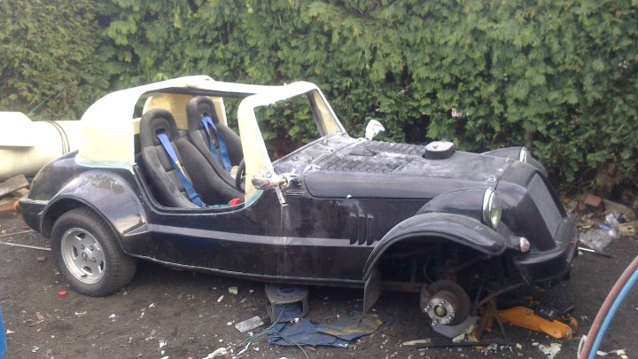 An old car is parked in a field - Pistonheads - The image depicts a small, old car that resembles a child's toy. It appears to be in poor condition, with parts missing and visible trash scattered near it. The car is colorful, painted in shades of black and yellow, and features a distinctive white hood. The vehicle's frame and parts from its interior suggest that it has been poorly maintained or abandoned. The car is positioned outdoors, likely on a driveway or street, as there is foliage and a fence visible in the background.