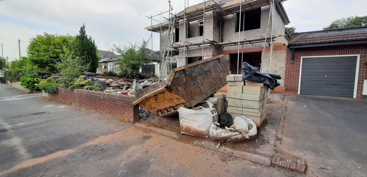 Skip lorry drivers - Page 1 - General Gassing - PistonHeads UK - The image shows a building under construction. The construction site is cordoned off with orange fencing, indicating ongoing work. There is a pile of rubble on the ground next to the building, which appears to be part of the renovation process. A brick wall can be seen in front of the construction site, possibly acting as a temporary barrier. In the background, there are other houses visible, suggesting that this construction is taking place in a residential area. The sky above is clear and blue, indicating favorable weather conditions for outdoor work.