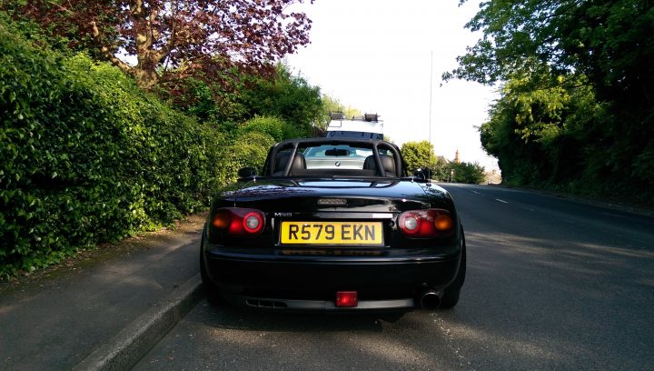 RE: Mazda MX-5: Catch It While You Can - Page 1 - General Gassing - PistonHeads - The image depicts a dark-colored, small sports car parked on a road beside a lush green hedge. The car's license plate is visible and reads "R579 EKN." In the background, the road winds through a shaded, wooded area with hints of sunlit trees. There is a white vehicle partially obscured by the hedge in the immediate background. The sunlight appears to be creating a dappled effect on the tree leaves.