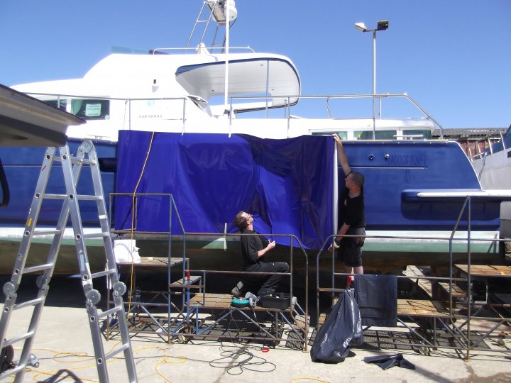 Wraps - Page 1 - Noble - PistonHeads - The image shows a marina scene where a blue tarp is being used to shield two individuals from the sun while they work on a white boat. The boat has its mast, or boom, raised and extends to the top of the frame, showing it to be a sailboat. The two people are wearing dark clothing and seem to be focused on a task. They are standing behind a metal railing, likely part of the dock or mooring area, which helps to secure the yacht. The setting suggests a typical marina environment.