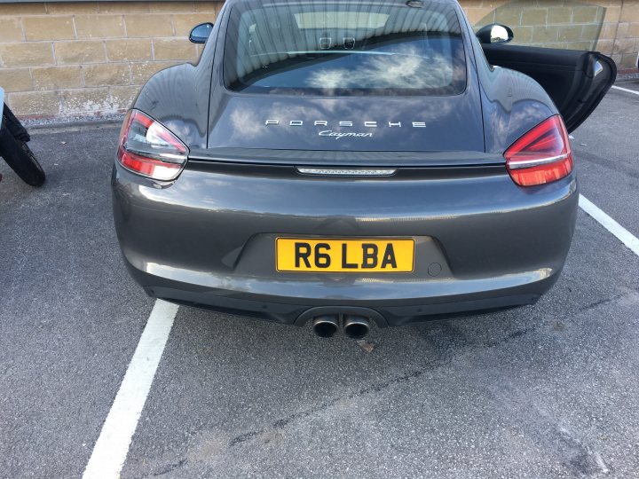 981 rear light upgrade - Page 1 - Boxster/Cayman - PistonHeads - In the image, there is a gray sports car parked in a mostly empty lot. The car is sleek with a gleaming finish, accentuated by the clear sky reflecting on its surface. The car is identified by its license plate, labeled with "RG6 LBA". The vehicle is a Porsche Cayman, as indicated by the model name near the license plate. The setting suggests a calm, possibly cool day, indicated by the open driver's side window.