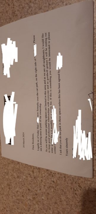 Residents parking purchased by local business - Page 1 - Homes, Gardens and DIY - PistonHeads UK - The image shows a piece of paper with a letter on it, sitting on a surface. The letter is written in English and includes multiple lines of text, but the specific content of the letter is not visible due to the blurred nature of the image. The background behind the paper appears to be an indoor setting, possibly a home or office environment. There's a partial view of a person's hand holding the paper from the bottom edge in the foreground, and it seems like the photo was taken by someone who is not visible in the frame.