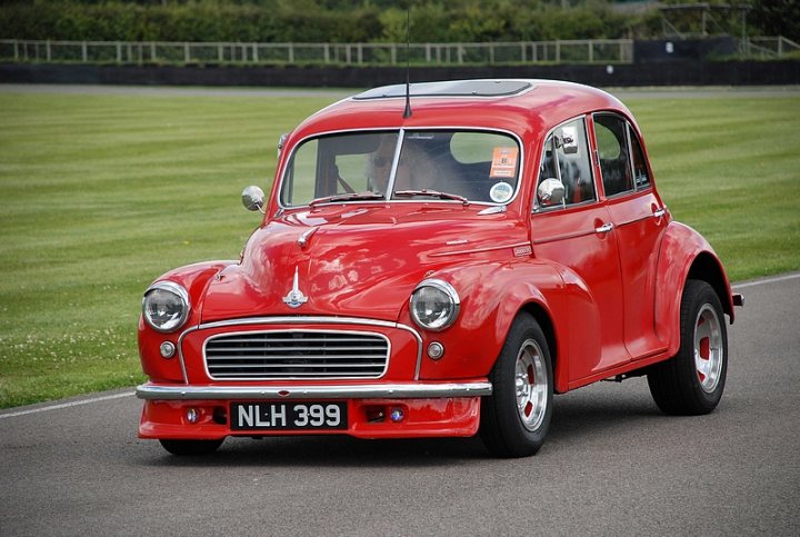 Goodwood Breakfast Club, Pre 1966 classic sunday - Page 1 - Goodwood Events - PistonHeads - The image features a vintage red car with a sunroof, driving on a paved road with colorful trees in the background. The car's license plate reads "NHL 399". There's a person visible through the car's windshield, but their features are not clear. The setting appears to be a sunny day due to the light reflecting on the car and the open sunroof. The car's bright red color stands out vividly against the greenery surrounding it.