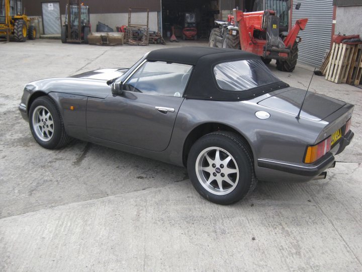 TVR23 V8S Body Off Restoration - Page 1 - S Series - PistonHeads - The image shows a black convertible car parked on a concrete surface. The car's top is down, revealing the interior. Its design features a body color that matches its roof and other parts of its body. Behind the car, partially visible, is a structure that appears to be an industrial or workshop building, which suggests a setting that might be a repair shop or a private parking space. The background is also populated with farming or construction equipment, indicating a commercial or agricultural setting. The overall scene conveys a sense of solitude, with the car being the main focus of the image.