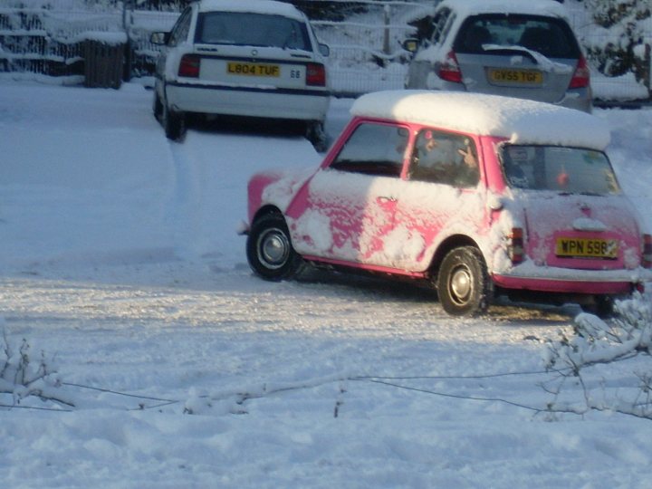 minis in the snow - Page 1 - Classic Minis - PistonHeads - This image captures a scene in an area blanketed with snow. In the foreground, there's a frozen, compacted layer of snow. A pink Miata car, coated in a thick layer of snow, is prominently positioned in the scene. Also snow-covered, further back, are three compact cars, each also in a parking lot. The cars are lined up facing one direction, indicating parked orientation. The setting appears serene, with the undisturbed white snow covering the landscape around the parked cars.