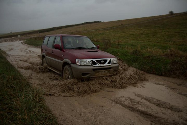 Is 4x4 shedding viable? - Page 1 - General Gassing - PistonHeads - The image shows a maroon SUV partially submerged in mud along an unpaved road. It seems to have gotten stuck, as evidenced by the mud surrounding the vehicle and covering the tires. The environment suggests a rural or remote area, as indicated by the absence of clear paths or structures that are typically found in the vicinity of roads. The sky appears overcast, indicating it might be raining or about to rain. There do not appear to be any visible people or animals in the scene.