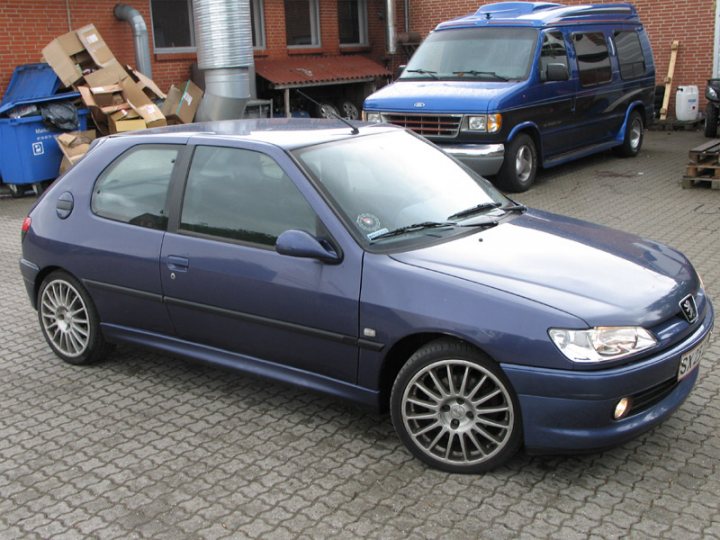 One owner FSH GTi-6 with 215K on the clock. Have I gone mad? - Page 1 - French Bred - PistonHeads - This image shows a blue car parked on a gray, textured surface. The car has five doors and is positioned against the backdrop of two other vehicles. The setting appears to be a parking area. To the left of the image, there's a commercial area with a blue dumpster containing assorted items, including a cardboard box. Additionally, red bricks can be seen in the background. There is a sense of depth from the middle of the car towards the horizon, suggesting the photo was taken from a perspective slightly behind the vehicle.