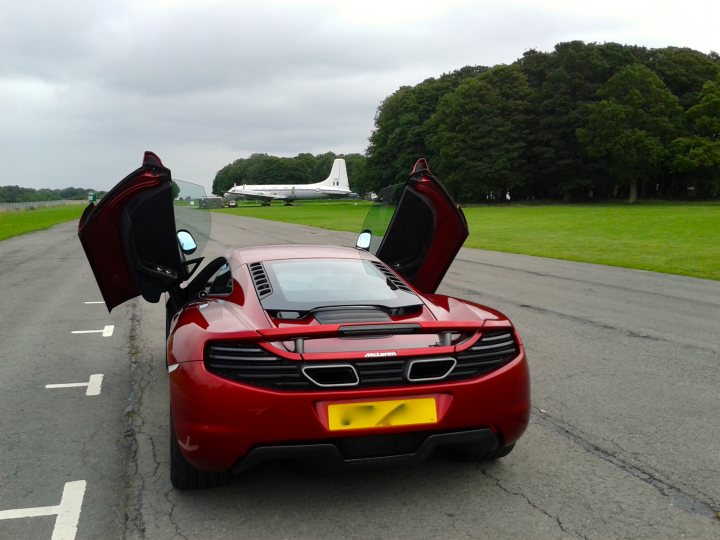 Finally - Page 1 - McLaren - PistonHeads - This image captures a scene at an airport. A luxury car, specifically a red McLaren, is parked at a terminal. The car's doors are open, inviting the viewer inside. The tarmac is visible behind the car, with a large airplane parked further back. The setting is outdoors, with ample natural light illuminating the scene. In the background, you can see a line of trees providing a serene backdrop to the bustling activity of the airport.